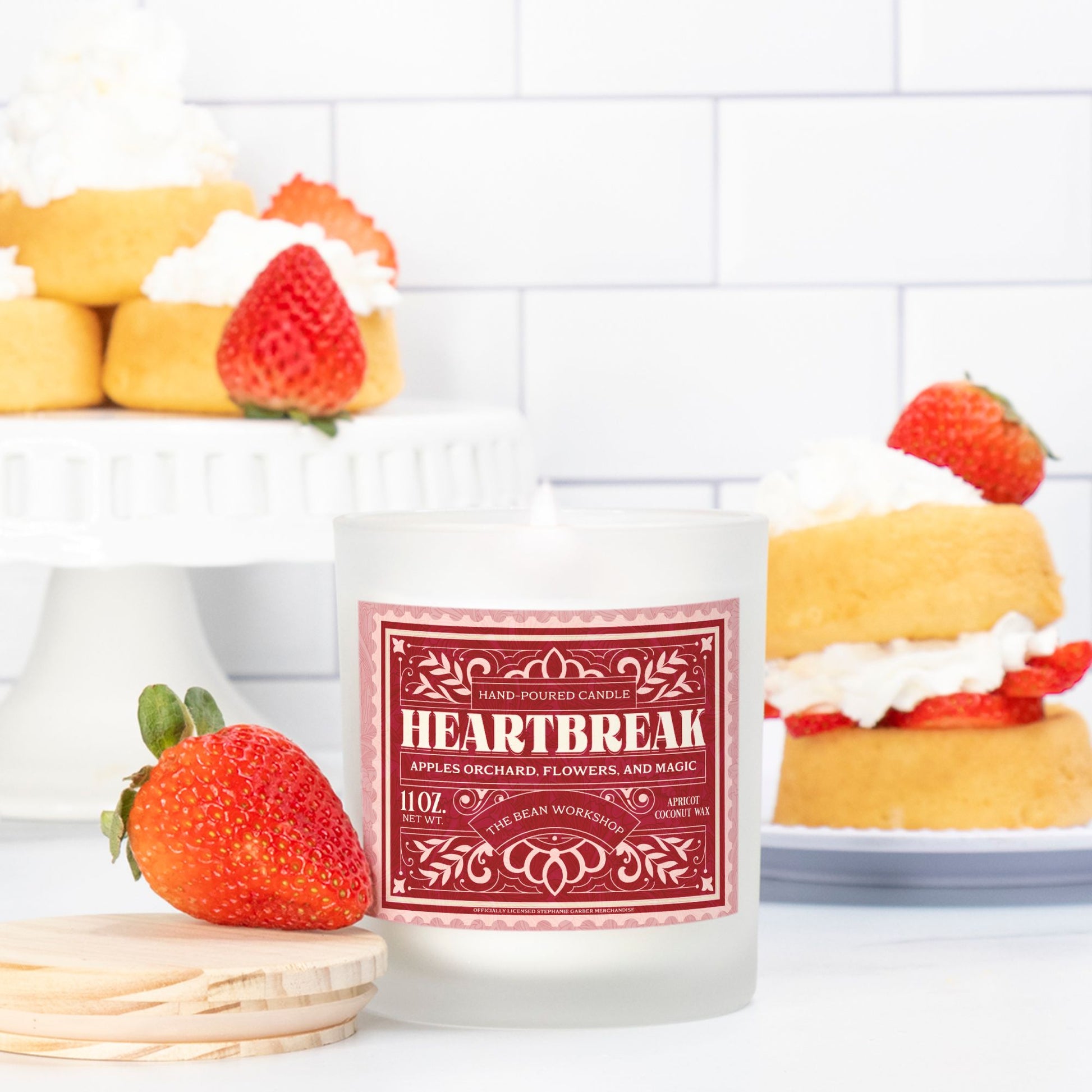 Heartbreak scented candle in frosted glass with pink label, surrounded by strawberries and shortcakes