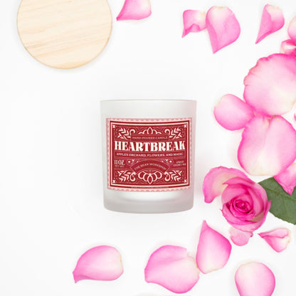 Heartbreak candle in frosted glass with pink wood wick, surrounded by pink rose petals.