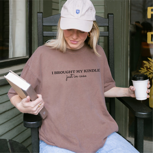 Woman wearing brown "I brought my Kindle just in case" shirt, holding book and coffee cup
