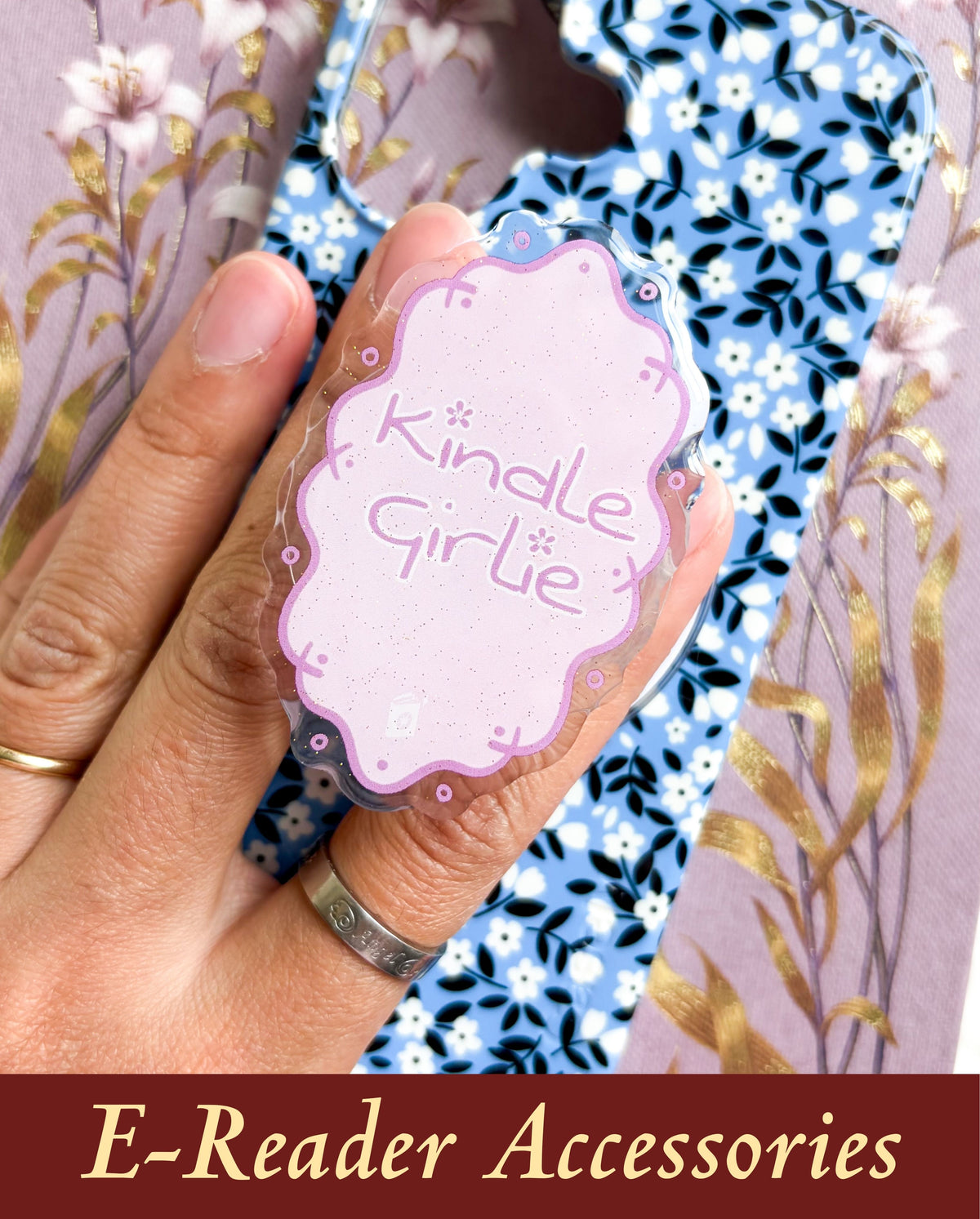 An aesthetic, lilac glitter Kindle Girlie e-reader accessory grip held over a patterned phone case, styled next to a gold-foiled book cover for a cozy, modern BookTok reading style layout.