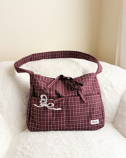 Large burgundy gingham book tote bag with multiple pockets and bow, displayed on white chair.