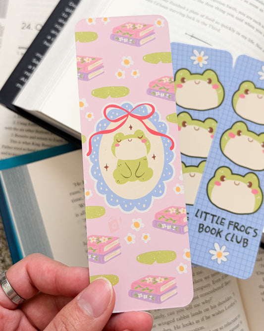 Hand holding a colorful frog-themed bookmark with more bookmarks in the background.
