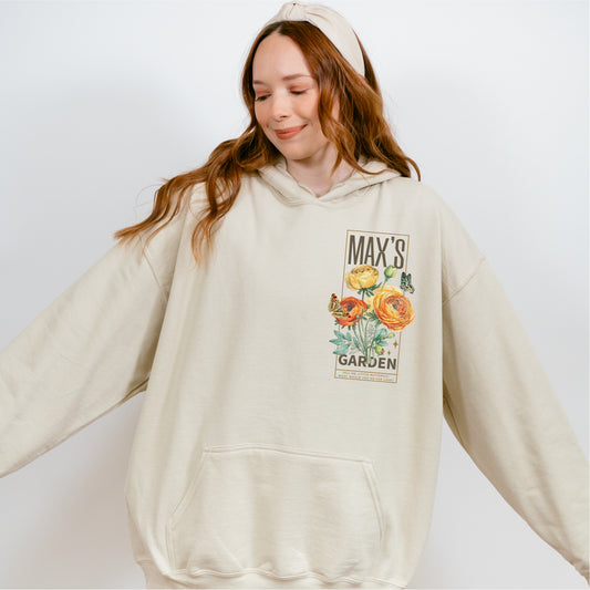 Woman wearing a cream hoodie with Max’s Garden floral graphic and butterflies on front