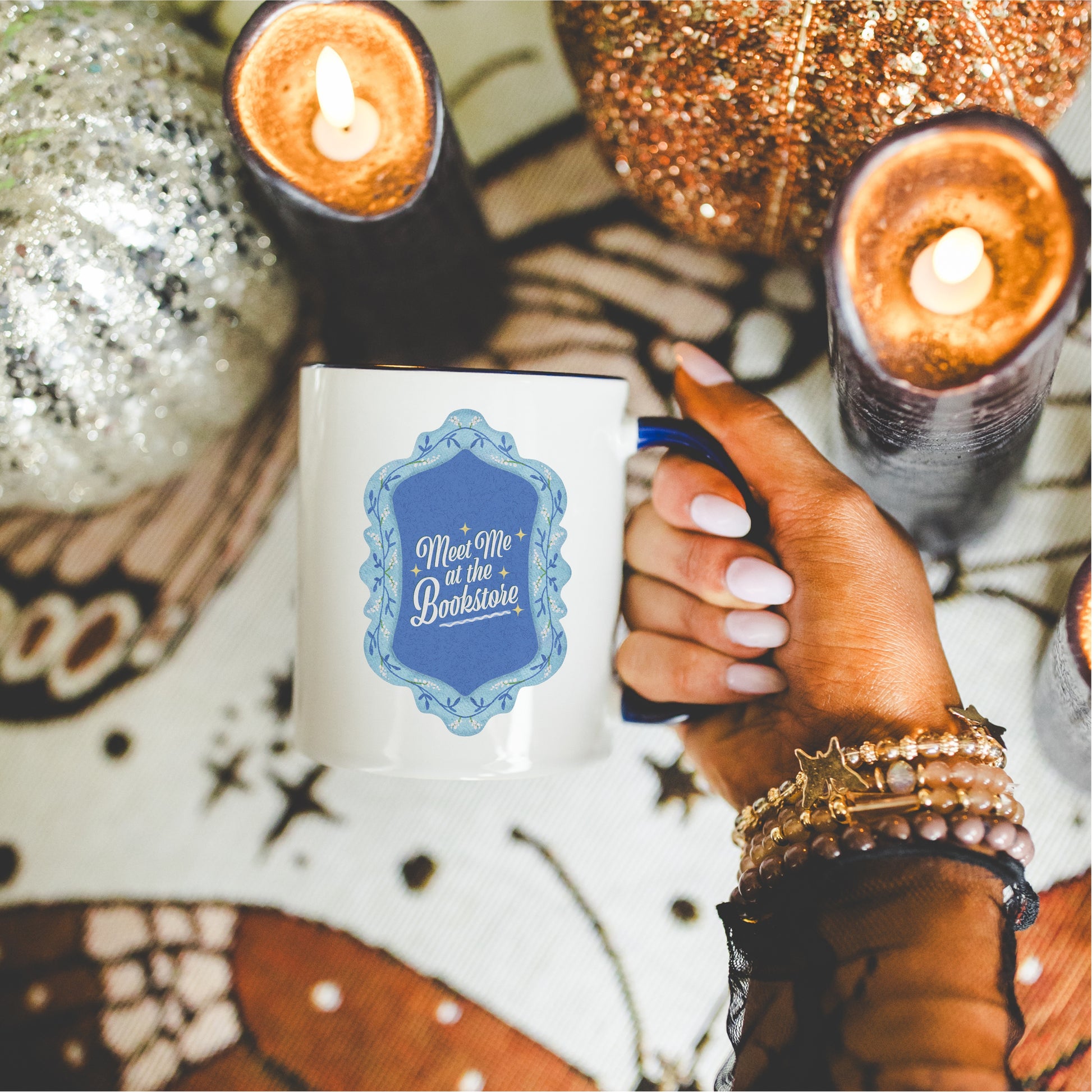 Hand holding 'Meet Me at the Bookstore' coffee mug, surrounded by candles and autumn decor