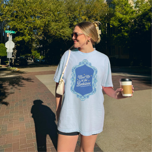 Woman in sunglasses wearing 'Meet Me at the Bookstore' graphic tee holding coffee outdoors