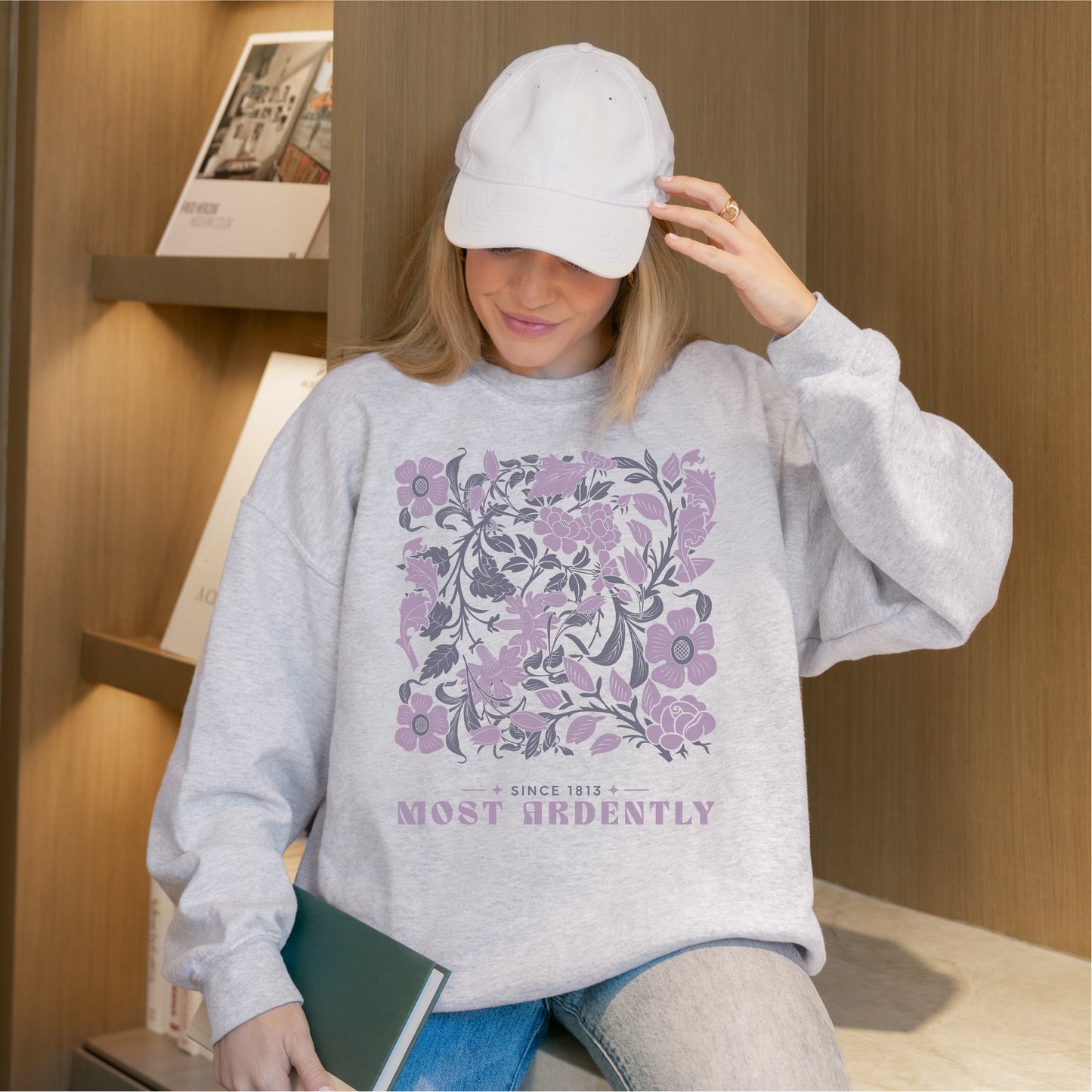 Woman wearing grey Most Ardently sweatshirt with purple floral design, holding a book