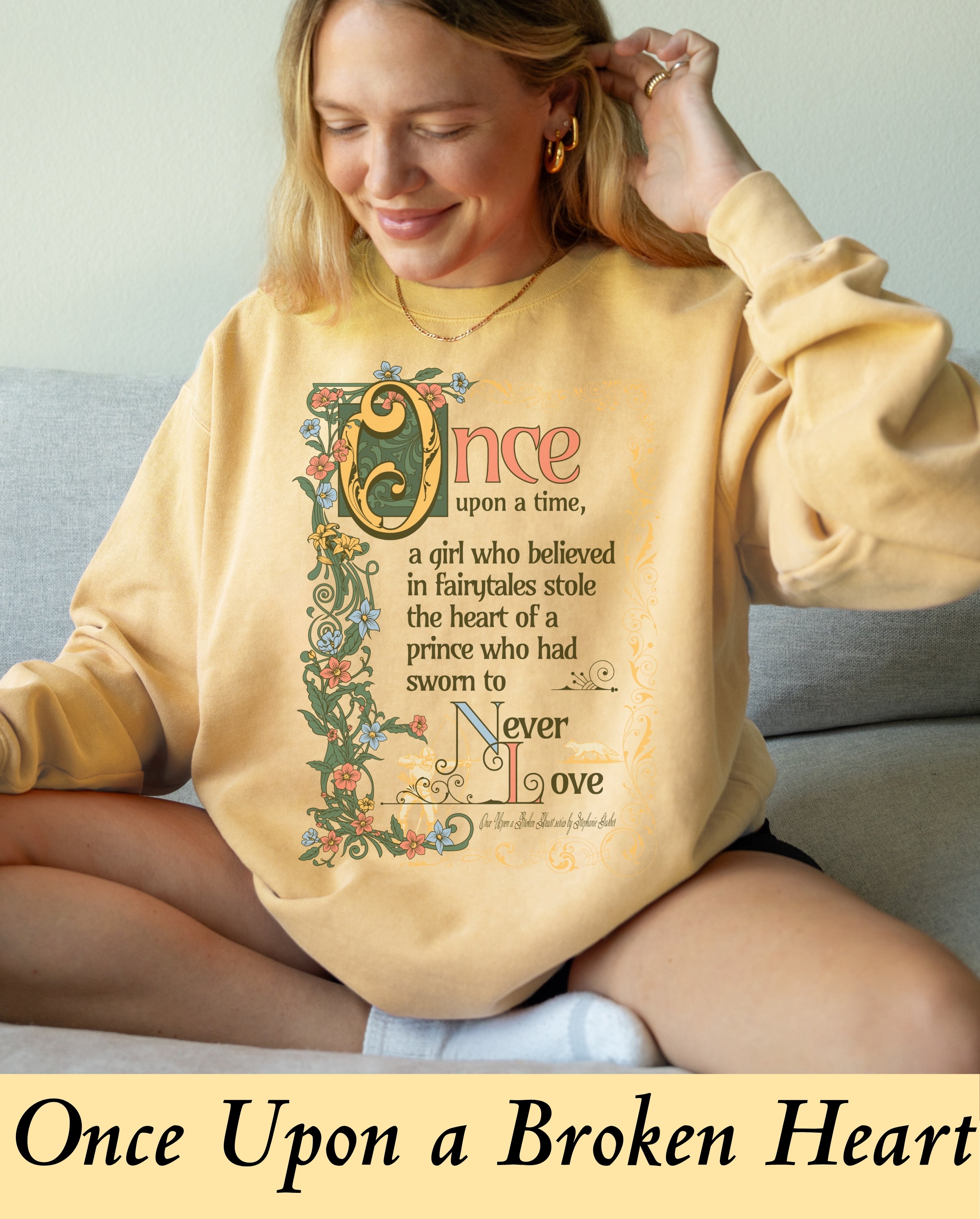 A model wearing an oversized, cozy butter-yellow crewneck sweatshirt featuring an illuminated fairytale manuscript style graphic. The text reads: 'Once upon a time, a girl who believed in fairytales stole the heart of a prince who had sworn to Never love,' adorned with an intricate floral border of vines, blue flowers, and orange blossoms, inspired by Stephanie Garber's Once Upon a Broken Heart.