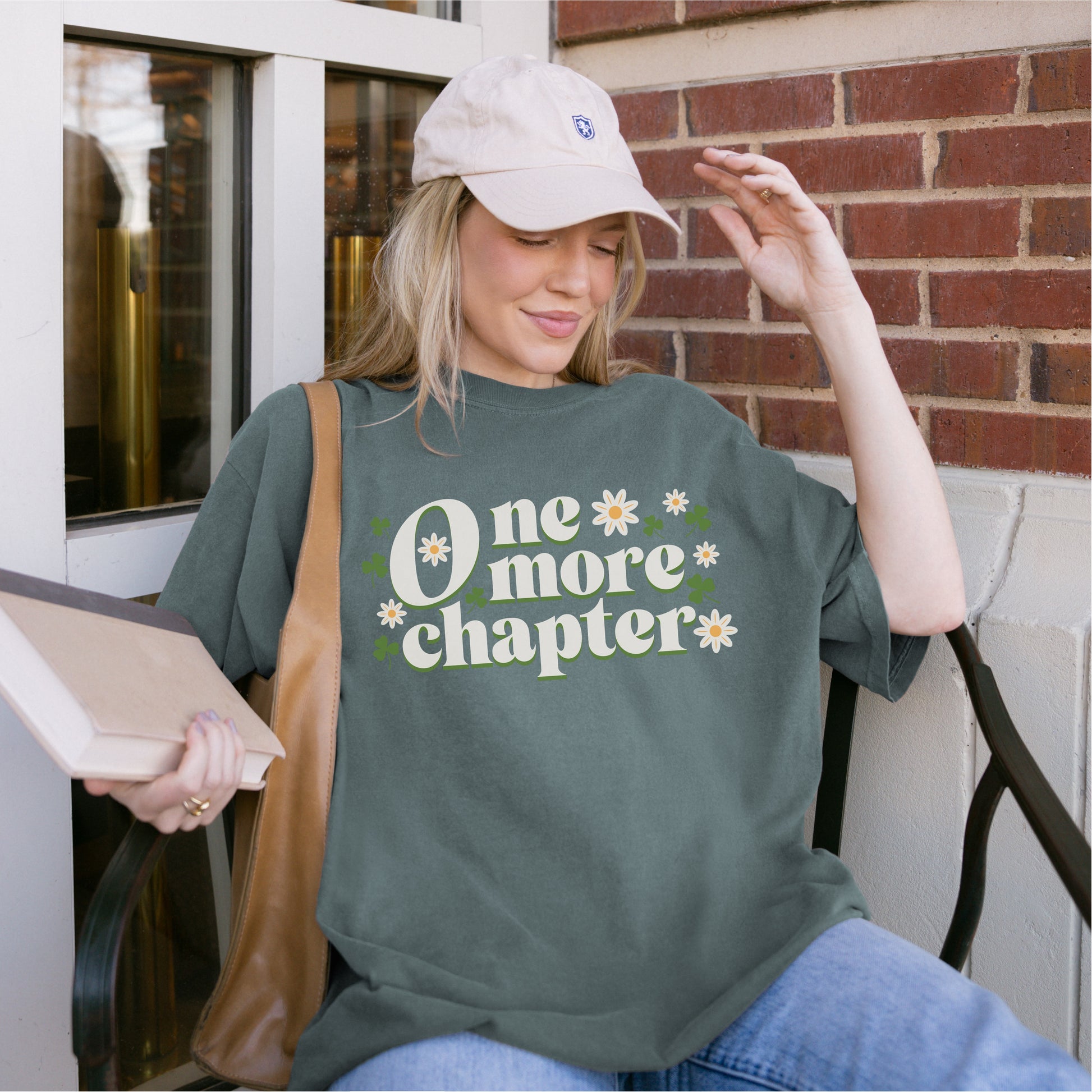 Woman in casual outfit holding book, wearing green 'One more chapter' book lover t-shirt with flowers