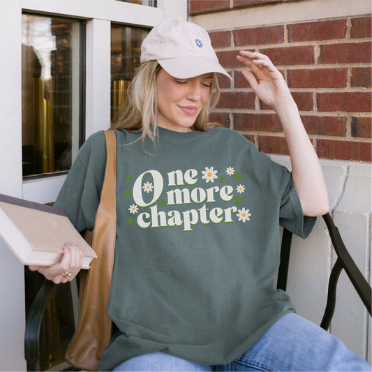 Woman in casual outfit holding book, wearing green 'One more chapter' book lover t-shirt with flowers