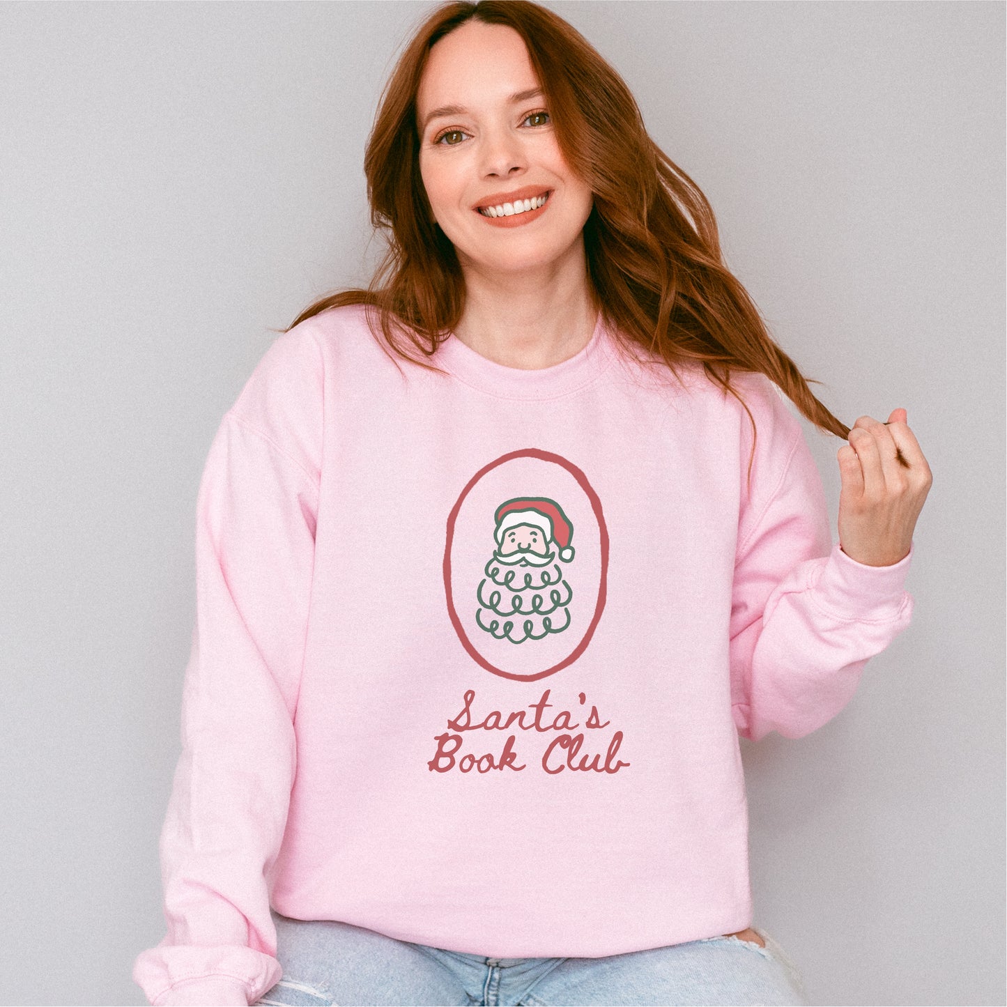 Woman wearing a pink Santa's Book Club sweatshirt with a Santa graphic and script text.