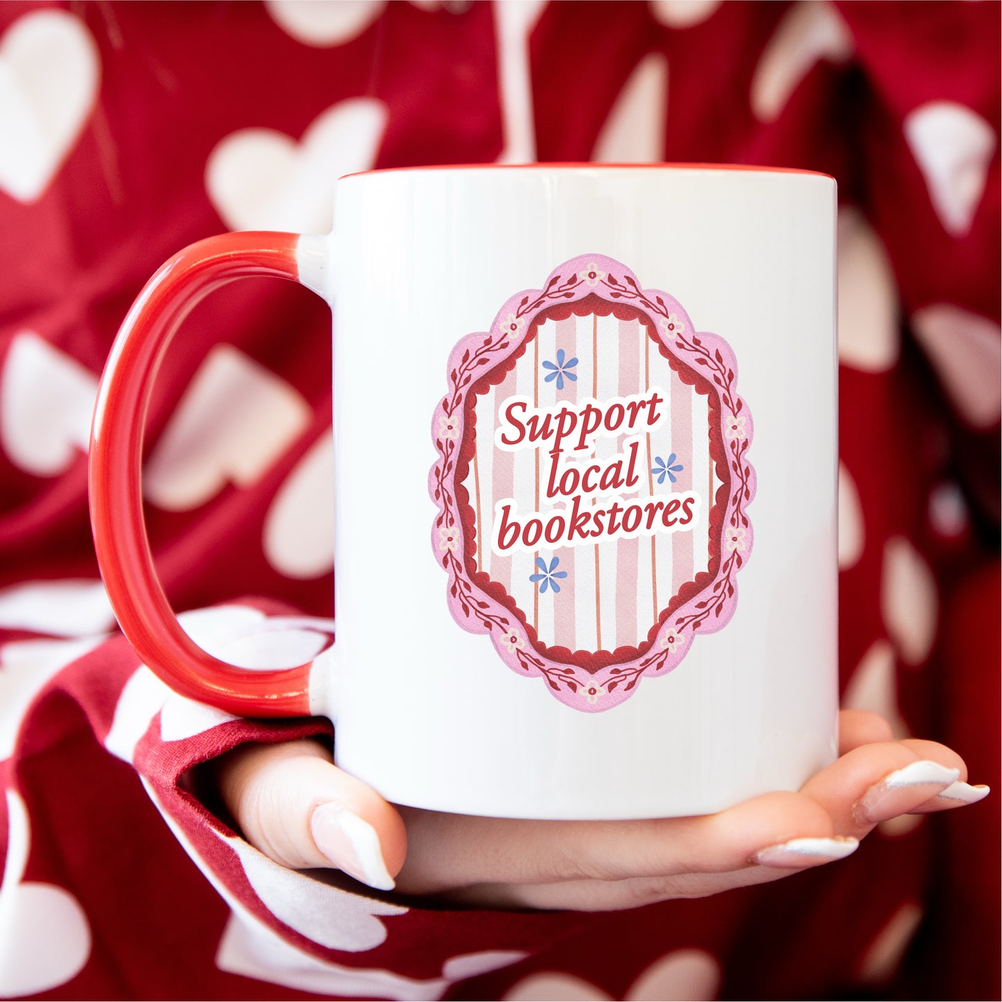 White and red mug with 'Support local bookstores' design, held by hand in polka dot pajamas