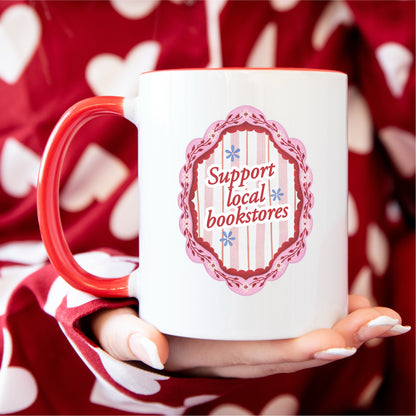 White and red mug with 'Support local bookstores' design, held by hand in polka dot pajamas