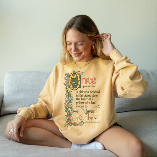 Woman wearing a yellow bookish sweatshirt with floral fairy tale quote graphic, sitting on a couch.