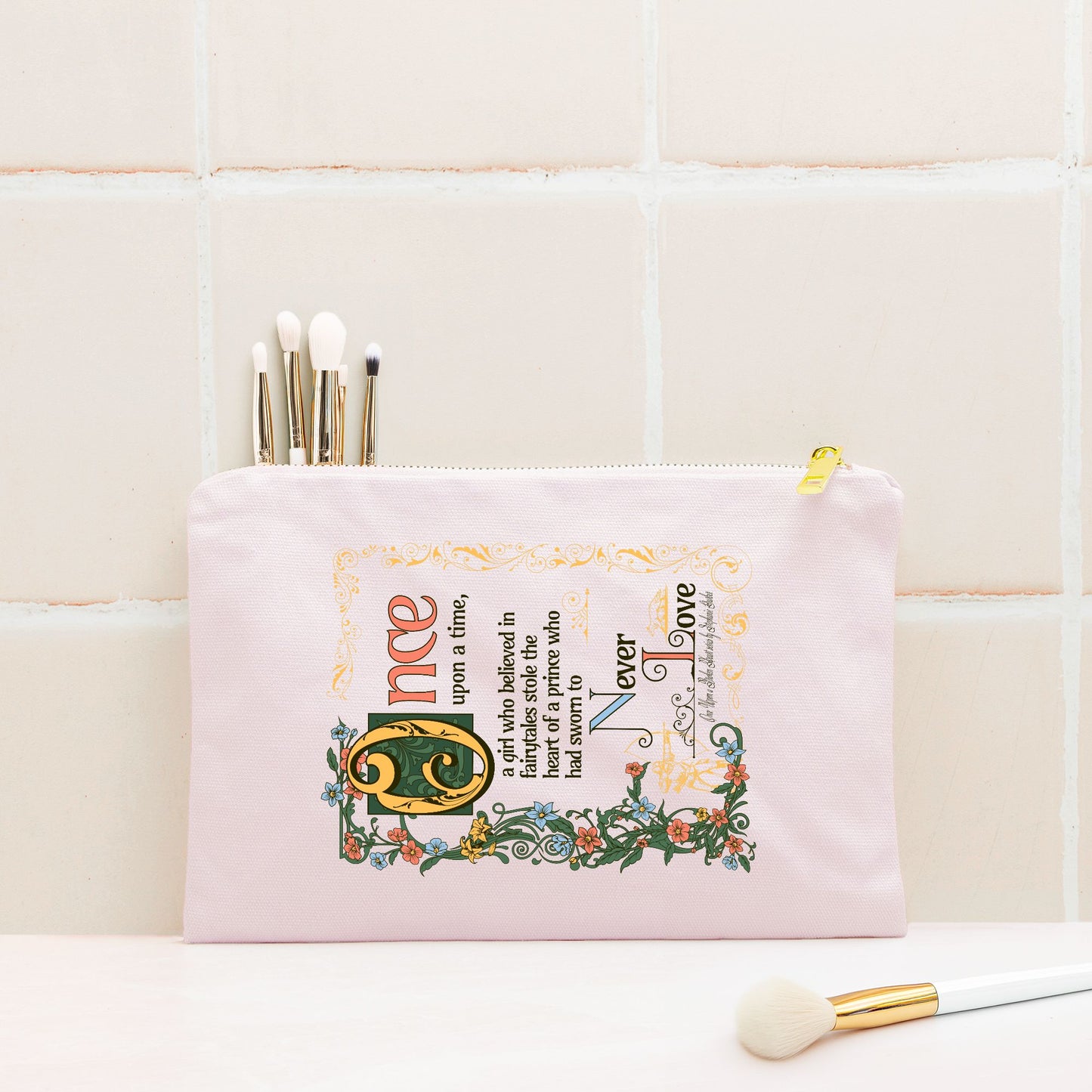 Pink illustrated zipper pouch with fairytale-inspired text and makeup brushes inside, on a tiled background.