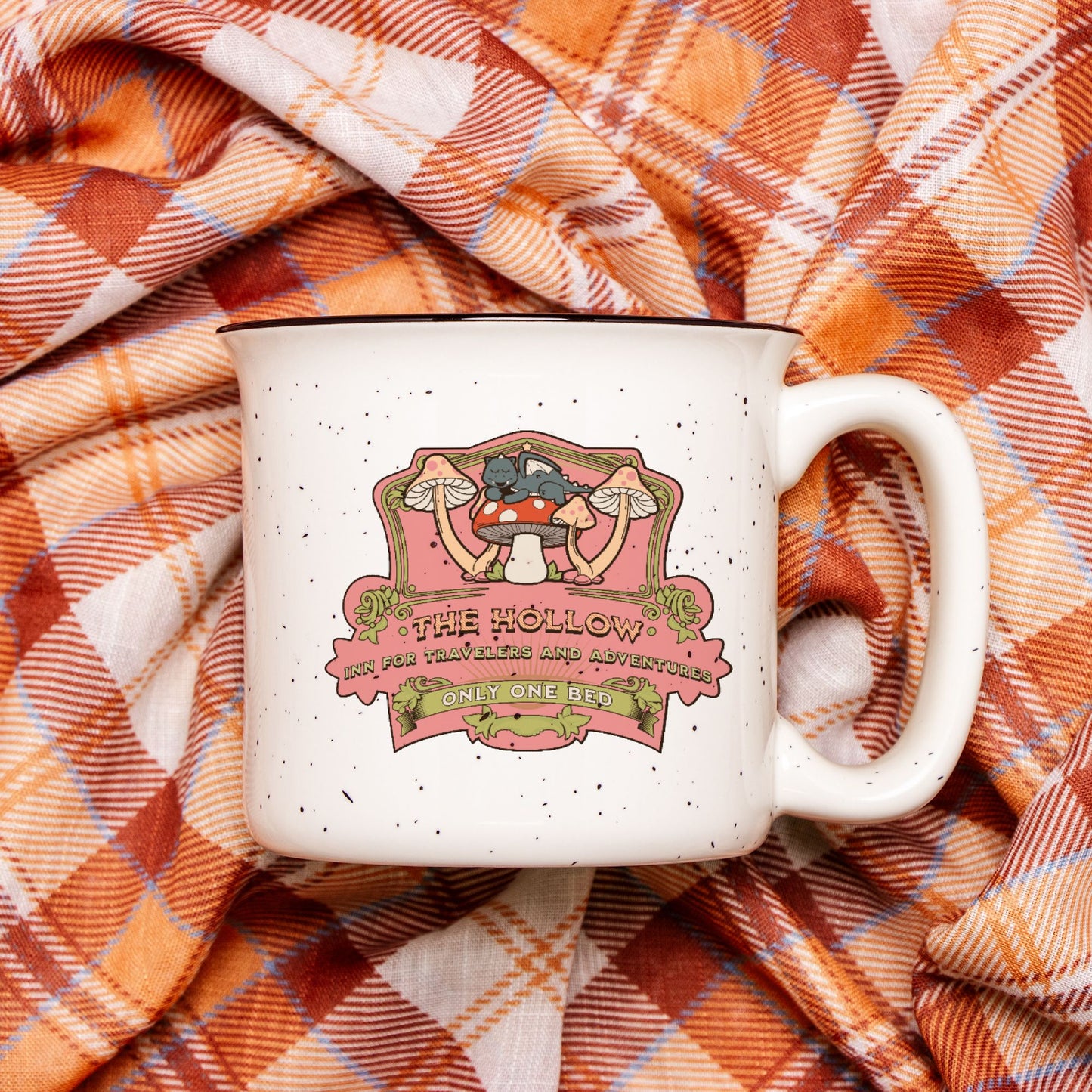 Speckled ceramic camp mug with whimsical mushroom illustration and bookish quote, on plaid blanket