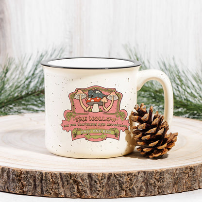 Speckled ceramic camp mug with whimsical mushroom illustration and bookish text, pinecone beside.