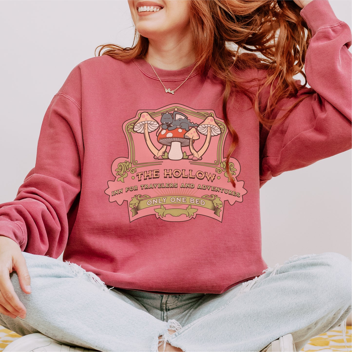 Woman in pink bookish sweatshirt with illustrated cats, mushrooms, and fantasy inn text