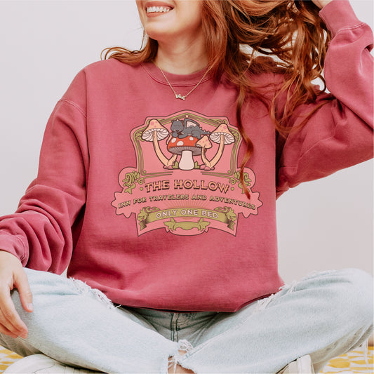 Woman in pink bookish sweatshirt with illustrated cats, mushrooms, and fantasy inn text