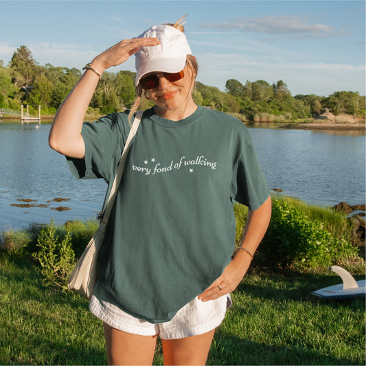 Woman outdoors by lake wearing green 'very fond of walking' bookish graphic tee and white hat