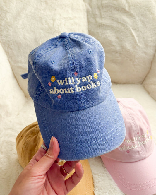 Blue denim cap with embroidered 'will yap about books' and floral accents, held by hand.