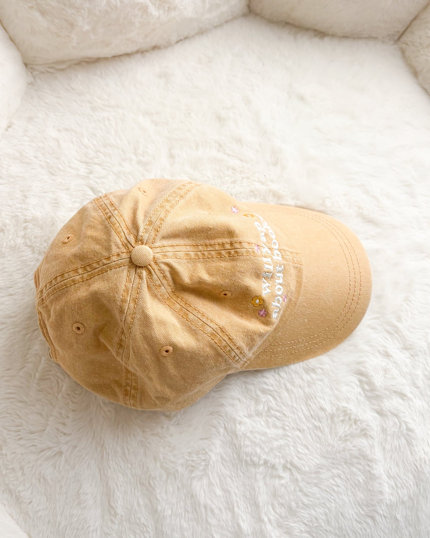 Tan embroidered baseball cap with book lover quote and small floral accents on white fluffy surface