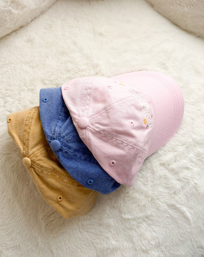 Three pastel baseball caps in pink, blue, and tan on a soft cream background