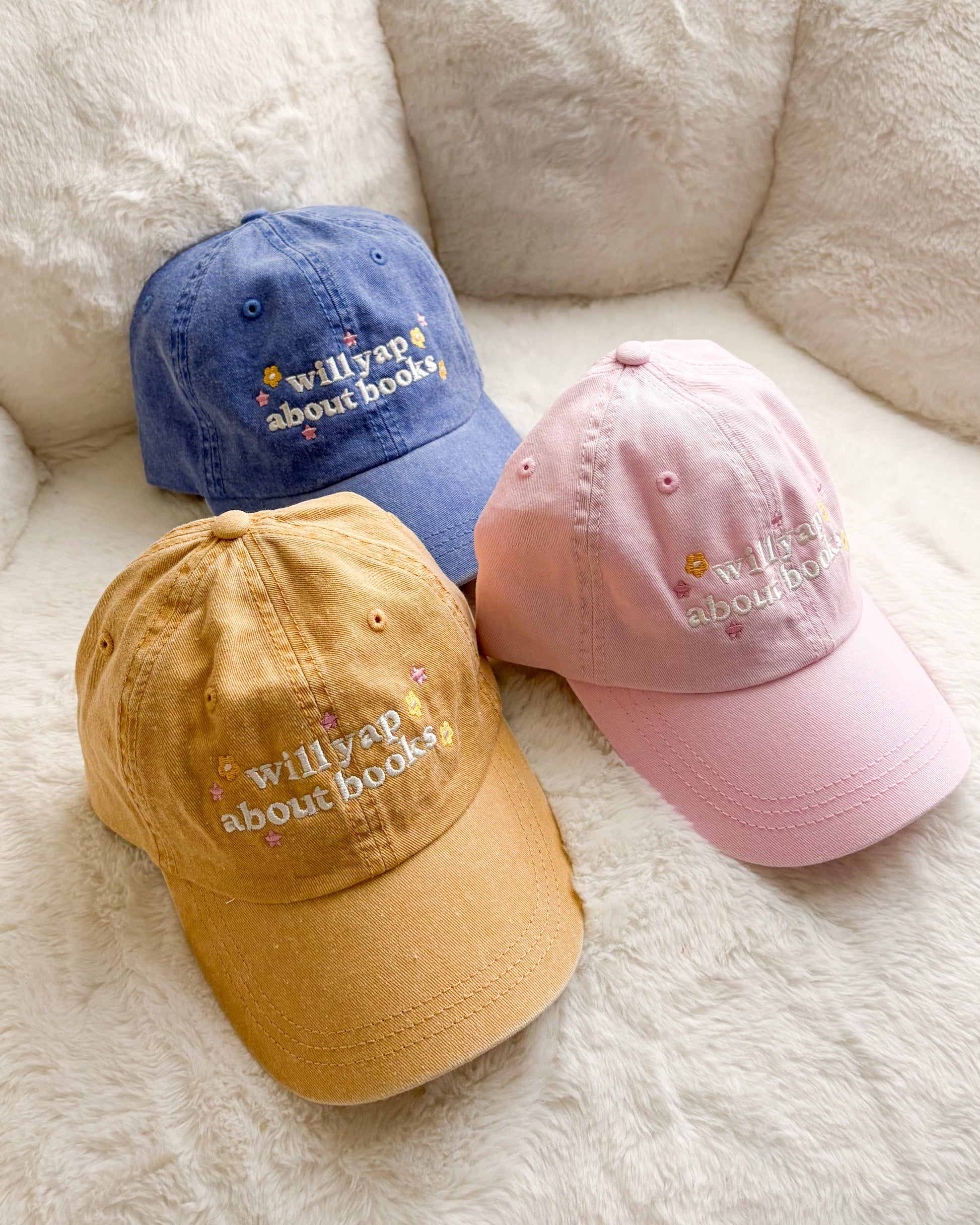 Three embroidered baseball caps in yellow, blue, and pink with 'willyap about books' text.