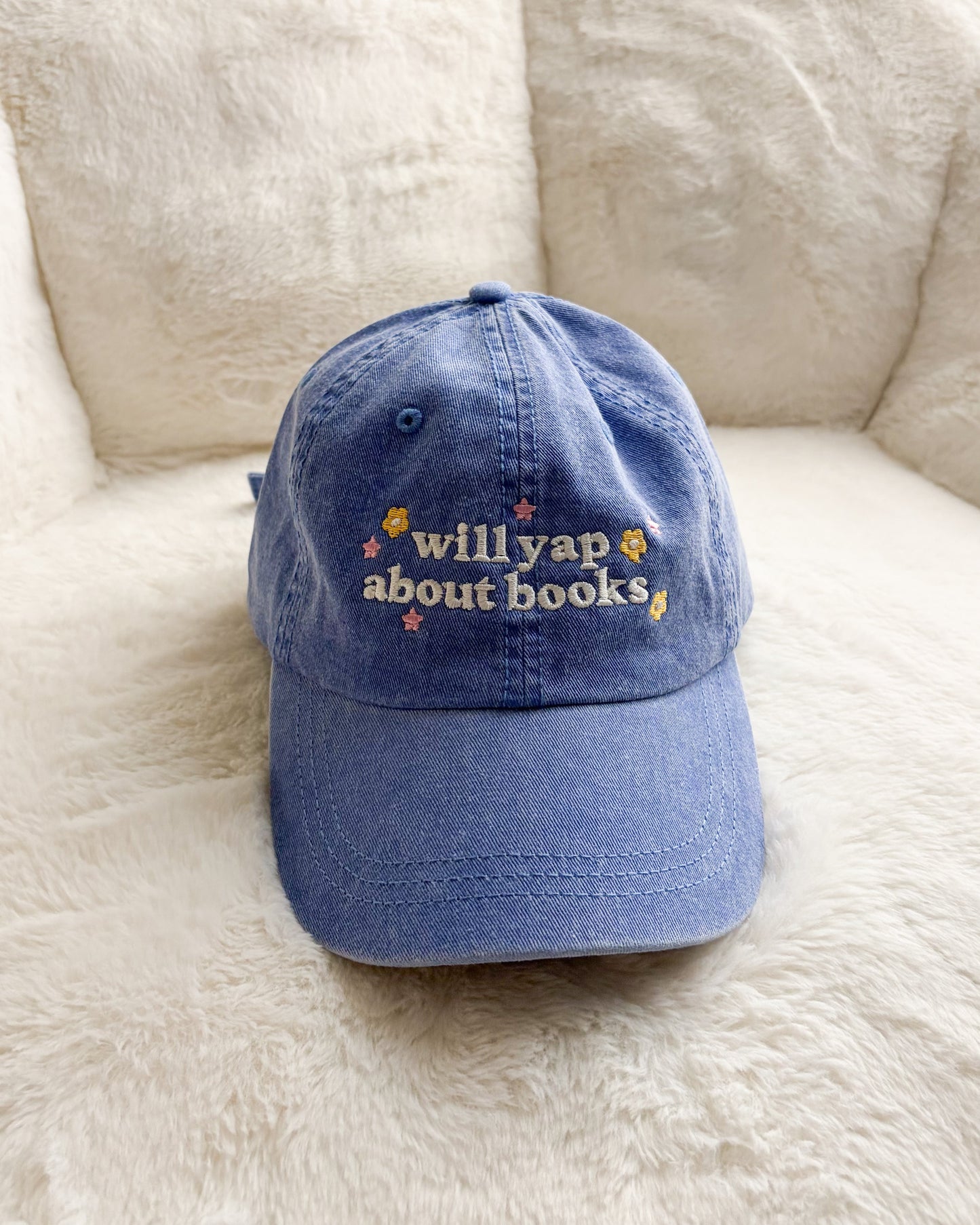 Blue embroidered baseball cap with 'will yap about books' text and floral designs on soft surface