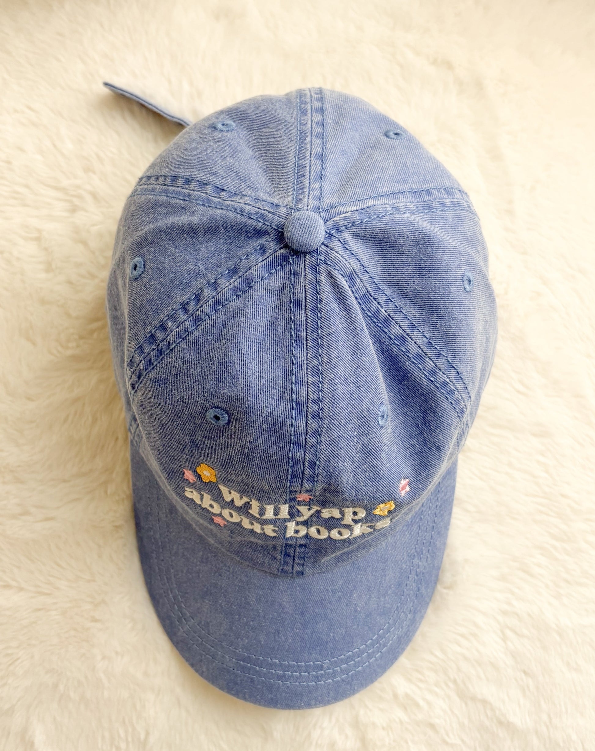 Blue denim cap with embroidered 'Will yap about books' text and small colorful flowers