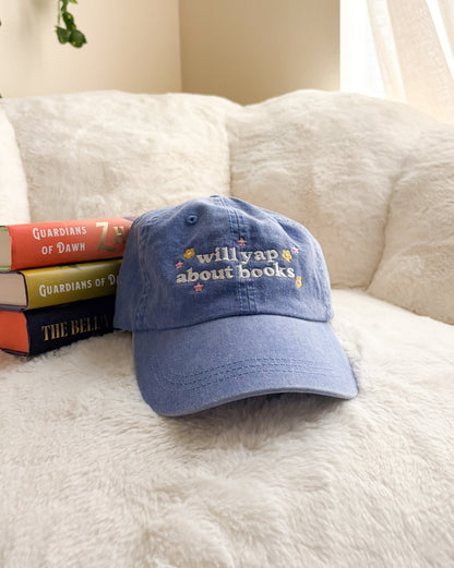 Blue embroidered 'will yap about books' hat with stacked fantasy books on a cozy chair