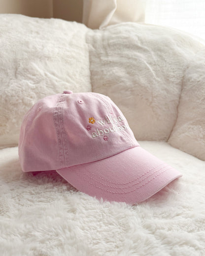 Pink embroidered baseball cap with 'write about books' text and book icon on a cozy white blanket