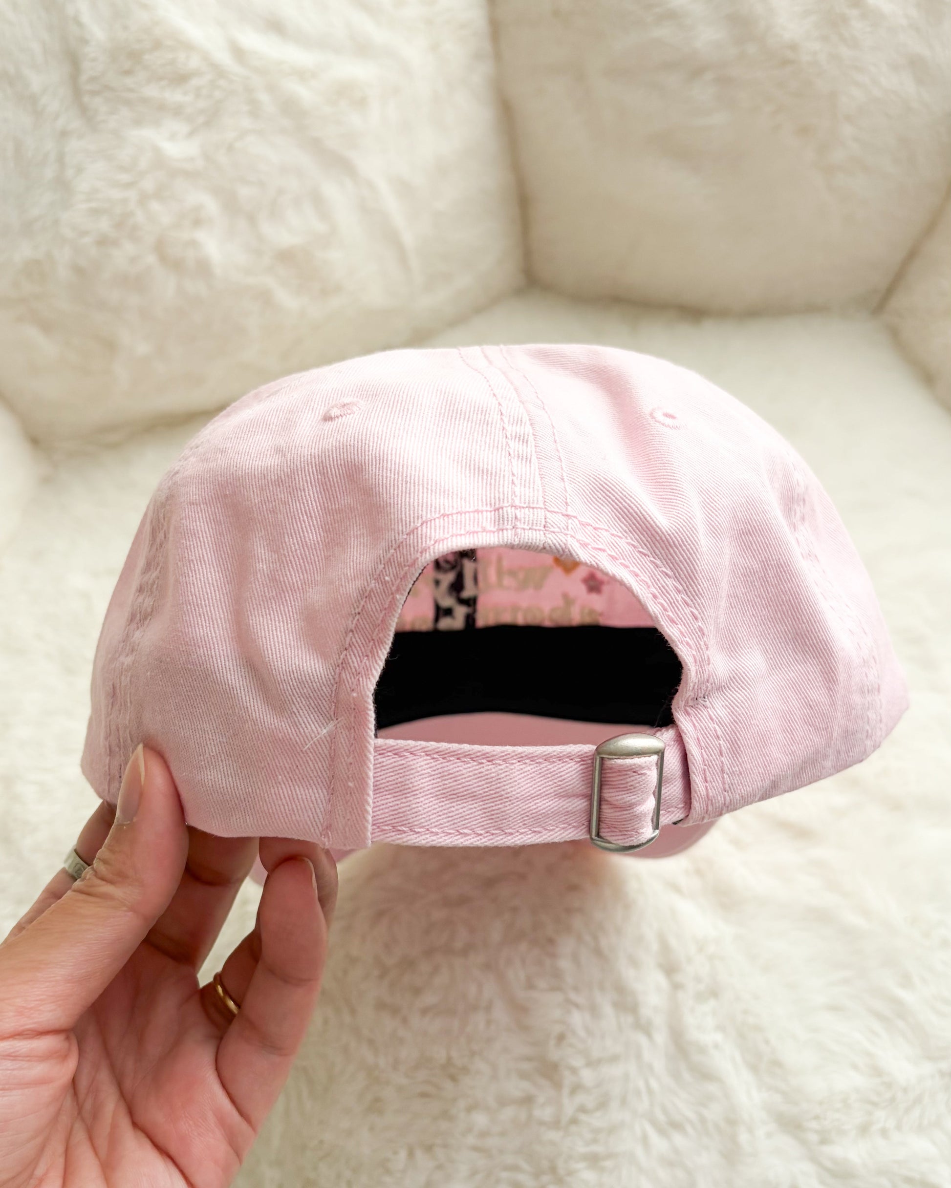 Hand holding pastel pink adjustable baseball cap with metal buckle, soft textured background