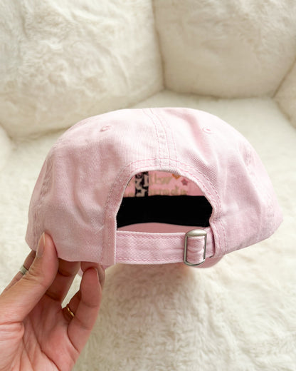 Hand holding pastel pink adjustable baseball cap with metal buckle, soft textured background