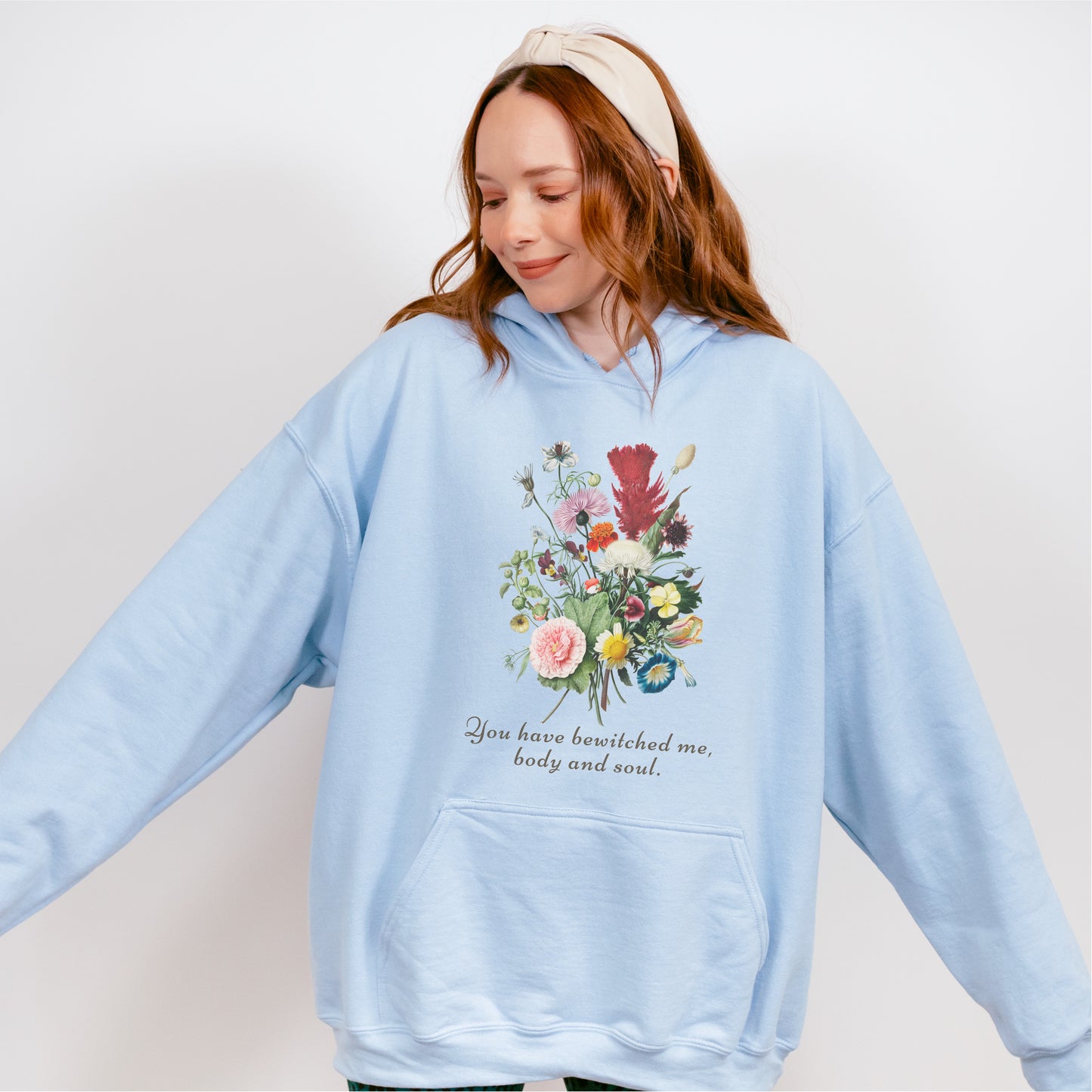 Woman wearing light blue hoodie with floral graphic and 'You have bewitched me, body and soul' text.