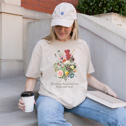 Woman wearing a cream tee with floral graphic and 'You have bewitched me, body and soul' quote, holding a coffee and book on outdoor steps.